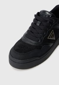 Baskets noires avec un dessus en daim et cuir texturé, présentant une semelle épaisse, des lacets noirs et un accent de logo métallique sur le côté.