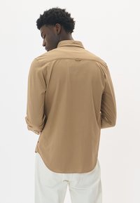 Svetlohnedá košeľa s dlhým rukávom a zadným yokeom, s golierom na gombíky a manžetami, skombinovaná s krémovými nohavicami. Hladká textúra.