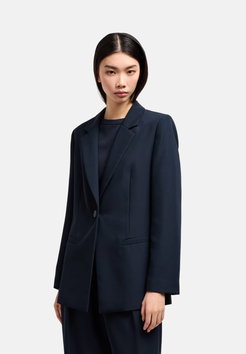 Blazer navy su misura con rever a punta, chiusura a un bottone e due tasche frontali. Tessuto liscio e vestibilità strutturata.