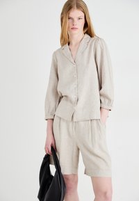 Camicia di lino beige a maniche corte, con bottoni sul davanti e vestibilità rilassata abbinata a pantaloni corti coordinati. Il modello tiene una borsa nera.