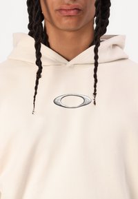 Creme-coloured hættetrøje med en forkert lomme; har et metallisk logo-detail. Blødt, struktureret stof med en justerbar snørerhætte.