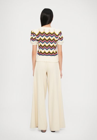 Pull en tricot à base crème, présentant des motifs zigzag multicolores. Associé à un pantalon beige à jambes larges, mettant en valeur la texture et le design.