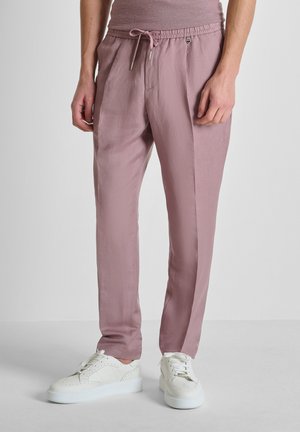 Persoon die mauve trekkoordbroek en witte sneakers draagt, staand tegen een effen lichte achtergrond.
