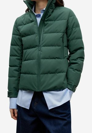 Mujer con chaqueta acolchada verde oscuro sobre una camisa azul claro y pantalones azul oscuro, con las manos parcialmente en los bolsillos.