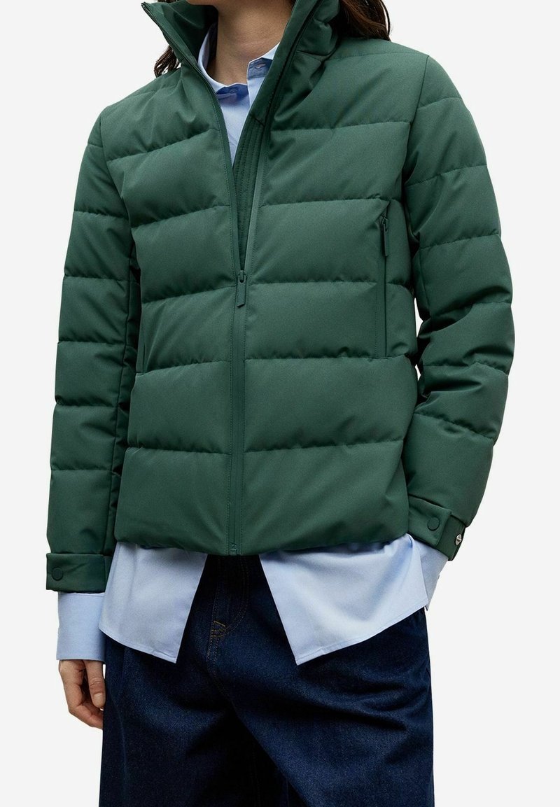 Femme portant une veste matelassée vert foncé par-dessus une chemise bleu clair et un pantalon bleu foncé, les mains partiellement dans les poches.