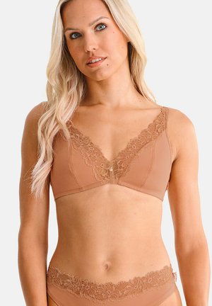 Vrouw draagt een bijpassend nude-kleurig kanten bh en ondergoed set, poserend tegen een eenvoudige lichte achtergrond.