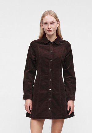 Jeune femme aux longs cheveux blonds portant une robe en velours côtelé marron foncé à boutons, manches longues et col.