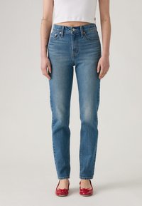 Μπλε τζιν παντελόνι με ψηλό μαξιλάρι, ίσια γραμμή, διακριτική φθορά και ραφές. Συνδυασμένο με κόκκινες φλατ παπούτσια και λευκή κοντή μπλούζα.