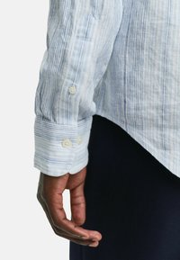 Camisa de rayas azul claro con puños largos y botones blancos, que presenta una textura suave y un corte relajado, combinada con una prenda oscura abajo.