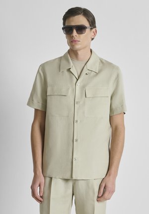 Jonge man draagt een licht beige kortärmelig overhemd met knopen en twee borstzakken en bijpassende broek, gecombineerd met donkere zonnebril.