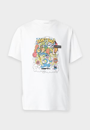 Mand iført en hvid t-shirt med farverige tegneserieprint, mørke vidde jeans og sorte sko, stående med hænderne i lommerne.
