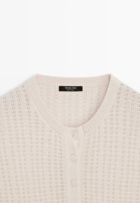 Cardigan tricoté rose clair avec un motif texturé, présentant un col rond, une fermeture par boutons et des bordures côtelées au niveau du col et de l'ourlet.