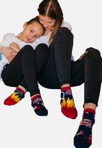 Calze rosse e blu navy con motivi di fiamme e veicoli. Una donna e un bambino siedono vicini, entrambi indossano pantaloni neri, con il bambino in una maglietta bianca.