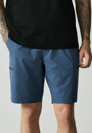 Muž nosi modré cargo kraťasy a čierne tričko, ukazuje tetované ruky a vrecká s zipsmi na kraťasoch.