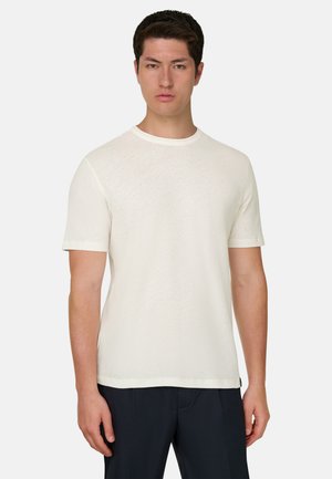 Modèle masculin portant un t-shirt blanc uni à manches courtes et col rond, accompagné d'un pantalon foncé, debout devant un fond clair uni.