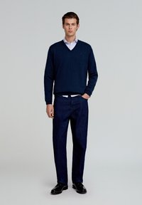 Pull en V marine sur une chemise à col bleu clair, assorti à un pantalon bleu foncé et des chaussures noires. Design simple et ajusté avec une texture lisse.