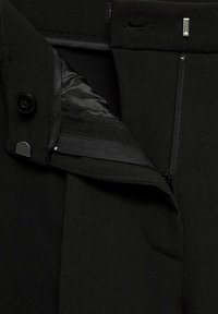 Pantalon noir en tissu lisse, avec fermeture éclair et bouton. L'intérieur présente une doublure élégante et une poche avec des détails de couture subtils.