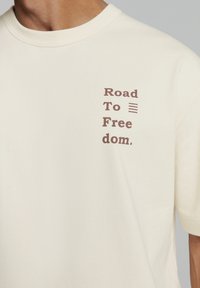 Crèmekleurig katoen T-shirt met bruine tekst: "Road To Free dom." geplaatst op de linkerkant van de borst in een moderne lettertype. Korte mouwen, klassieke ronde hals.