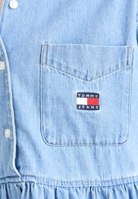 Denim haljina svijetloplave boje s kopčanjem sprijeda, odgovarajućim gumbima i džepom na prsima s crvenim, bijelim i plavim logom "TOMMY JEANS".