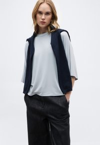 Νεαρή γυναίκα με ανοιχτό μπλε μπλουζάκι σε μεγάλο μέγεθος, μαύρο παντελόνι με φαρδιά πόδια και μπλε σκούρο πουλόβερ κρεμασμένο στους ώμους, μπροστά σε απλό φόντο.