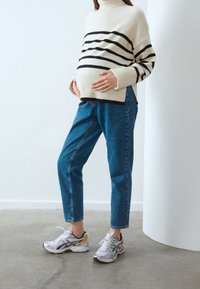 Randig krämfärgad och svart höghalset tröja, blå högmidjade jeans och silverfärgade löparskor med detaljer. Modellen håller om en gravid mage.