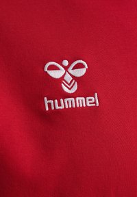 Czerwona tkanina sportowa z haftowanym białym logo "hummel" oraz abstrakcyjnymi kształtami i liniami. Gładka struktura o dopasowanym kroju.