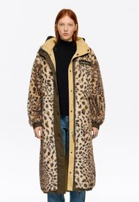 Mujer con cabello rojo que lleva un abrigo largo de piel sintética con estampado de leopardo sobre un suéter negro y jeans azules, de pie frente a un fondo blanco.