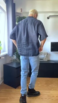 Homem com cabelo loiro curto, vestindo uma camisa escura larga, jeans azuis claros e botas pretas, de pé em uma sala clara com uma máquina de costura e plantas.