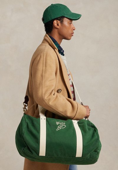 Νεαρός άνδρας με γκρι ζακέτα και πράσινο καπέλο, που κρατάει μια πράσινη τσάντα duffel Polo με λευκές τιράντες ώμου, στραμμένος προς τα δεξιά.