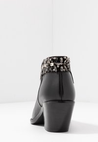 Bota de cuero negra hasta el tobillo con un tacón grueso y adornos de cuentas decorativas alrededor del cuello. Textura suave con un acabado pulido.