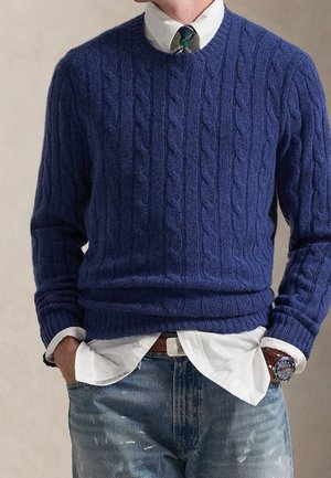 Homme portant un pull en maille torsadée bleu sur une chemise blanche avec une cravate à carreaux verts et bleus, associé à un jean bleu déchiré et une montre-bracelet.