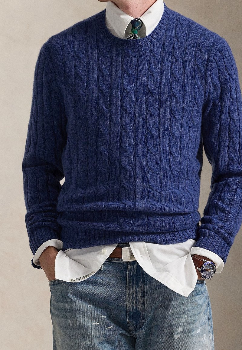 Homme portant un pull en maille torsadée bleu sur une chemise blanche avec une cravate à carreaux verts et bleus, associé à un jean bleu déchiré et une montre-bracelet.