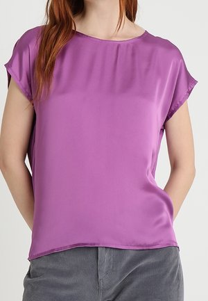 Mujer con blusa de satén morada de manga corta y holgada, combinada con pantalones grises, de pie con las manos en los bolsillos.