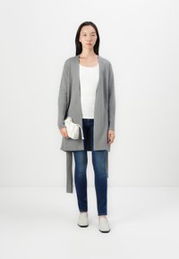 Szary sweter dzianinowy z otwartym przodem i długimi rękawami, noszony na białej koszuli i niebieskich dżinsach. Trzyma biały worek ze sznurkiem. Gładka tekstura.