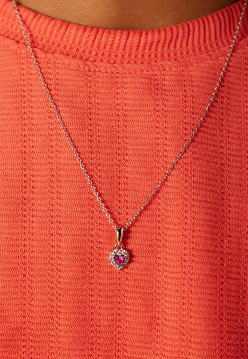 Zilveren ketting met een hartvormige hanger met een centraal roze edelsteen, omringd door kleine heldere steentjes, gedragen over een textuur koraalkleurige top.