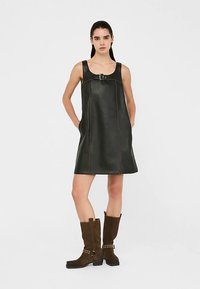 Schwarzes Lederkleid in A-Linie mit breiten Trägern, vorne mit einer Schnallendetail und sichtbaren Nahtnähten. Das Model kombinierte es mit braunen Stiefeln.