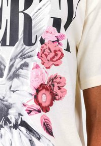 Beige t-shirt met korte mouwen, voorzien van grote zwarte letters, bloemenontwerpen in roze en rood en een gestructureerde grafische print van een adelaar.