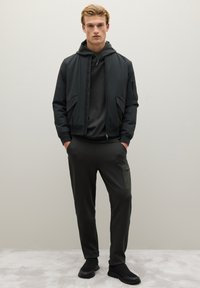 Zwarte bomberjack over een donkere hoodie, gecombineerd met donkere joggers met een groene zak. Matte, gladde stoffen; casual atletische stijl.
