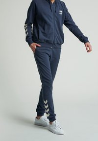 Námořnicky modrá mikina s kapucí a tepláky z měkké látky. Má bílé šipkové proužky na rukávech a manžetách kalhot, s logem na hrudi. Nosit se bílé tenisky.