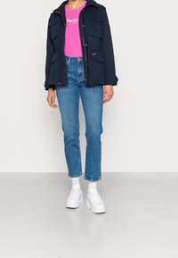 Granatowa kurtka z wieloma kieszeniami, różowa koszulka z białym logo, niebieskie dżinsy i białe sneakersy na platformie, wszystko na neutralnym tle.