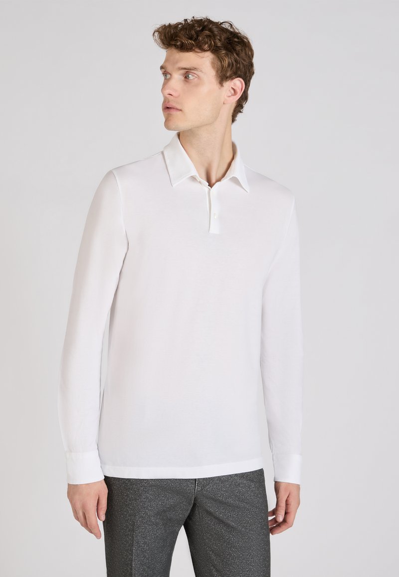 Witte poloshirt met lange mouwen, gekenmerkt door een klassieke kraag en drie knopen aan de voorkant. De stof is glad met een getailleerde pasvorm. Draagt een combinatie met grijze textuurbroek.
