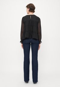 Frau in einer schwarzen transparenten Bluse mit langen Ärmeln und gepunktetem Muster, kombiniert mit dunkelblauen engen Jeans, die mit dem Rücken zur Kamera auf einem einfarbigen Hintergrund steht.