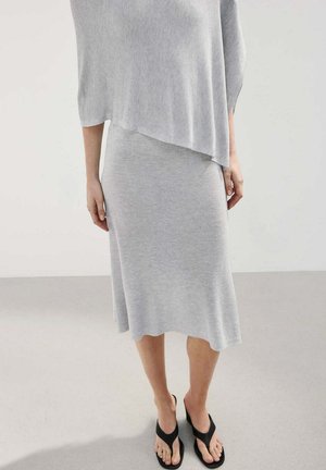 Mujer con falda midi de punto gris claro y top asimétrico a juego con sandalias negras tipo chancla, de pie sobre un suelo gris.