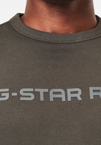 Sudadera verde oscura con cuello redondo y bordes acanalados. Presenta una marca en gris claro y en negrita en la parte frontal. Textura de tela suave.