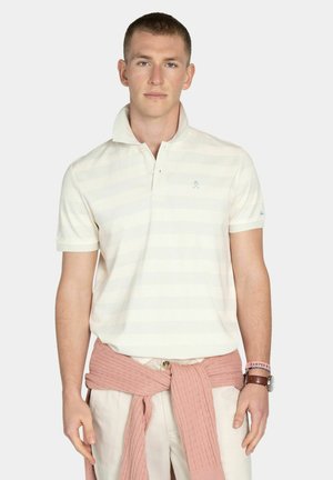 Młody mężczyzna w białej koszulce polo w paski, beżowych spodniach i różowym swetrze związanym wokół talii, z zegarkiem i bransoletkami na nadgarstkach.