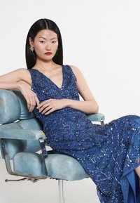 Modrá večerní toaleta s flitry, s výstřihem do V, přiléhavým střihem a složitými vzory korálků. Model sedící na vintage modré židli, která vystihuje eleganci.