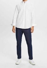 Weißes Langarm-Hemd mit Knopfleiste und Kragen, kombiniert mit dunkelblauen Jeans und weißen Sneakers, vor einem neutralen Hintergrund stehend.