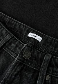 Jeans de mezclilla negros con cierre de botón, cinturilla cosida y una etiqueta que dice "TWO SOON" con talla 31/32. Textura sutil visible.