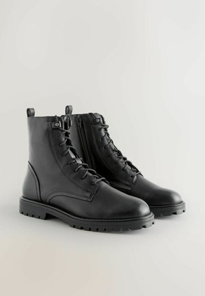 Mustat nahkaiset nilkkasaappaat kengännauhoilla, sivuvetoketjulla, kumipohjalla ja teksturoidulla ompeleella. Takana on lenkki, joka helpottaa pukemista.