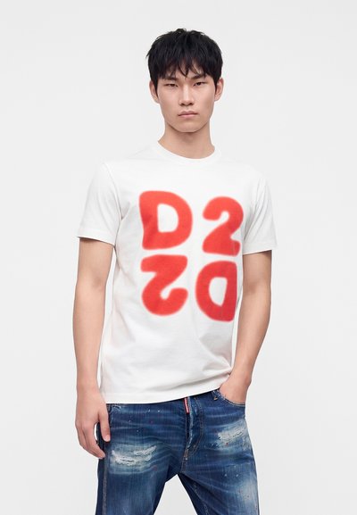 Jeune homme debout, une main dans la poche, portant un t-shirt blanc avec de grandes lettres rouges stylisées D2 et un jean bleu délavé.